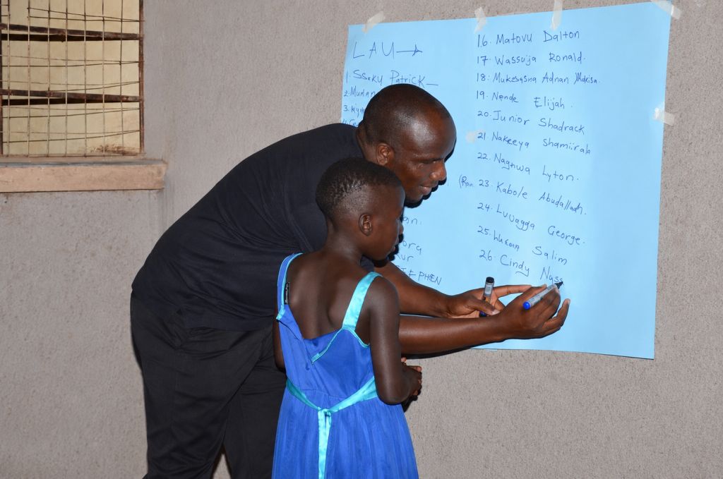 Diphas Kiguli, the Board Vice Chairperson of Literacy Aid Uganda helping one of the children on how to write her name